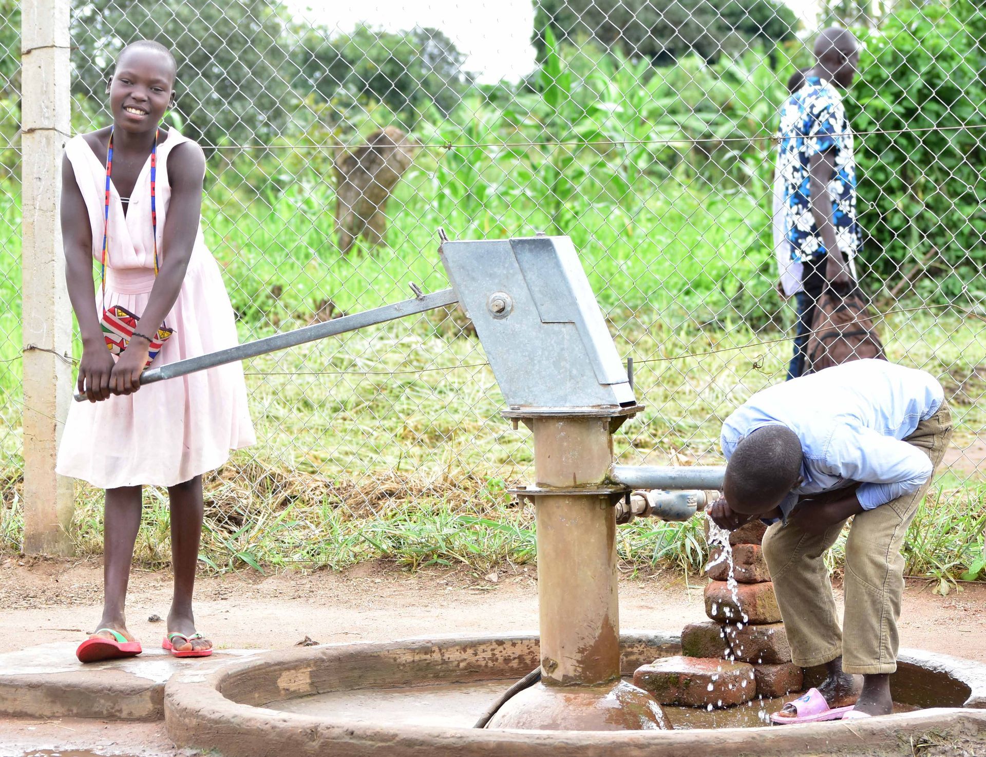 A girl in a pink dress is standing next to a man drinking water from a well.