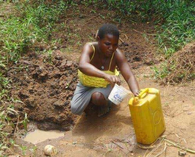 Person crouches by a muddy stream, filling a yellow jerrycan with water.