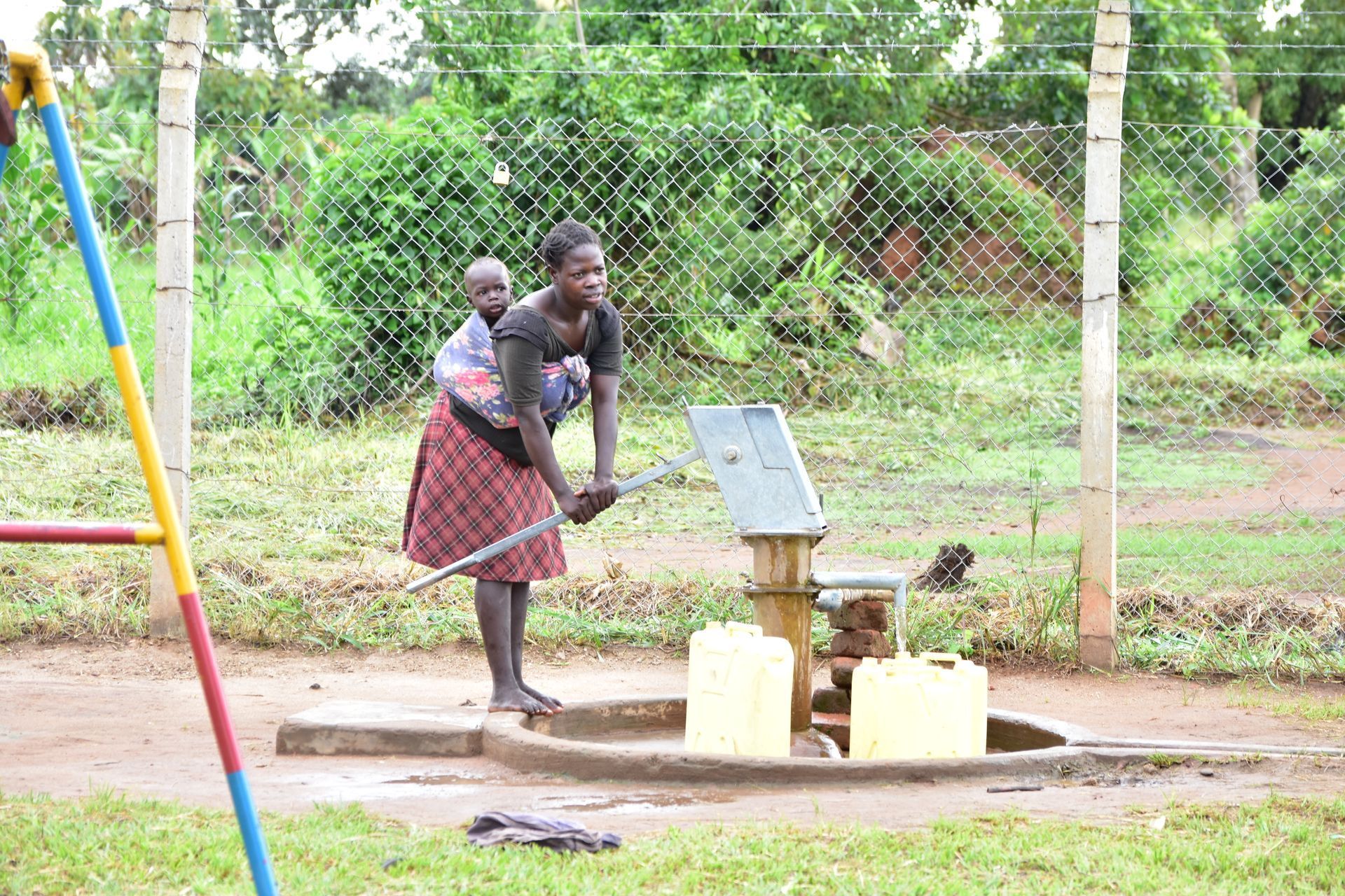 A woman is carrying a baby on her back near a water pump.