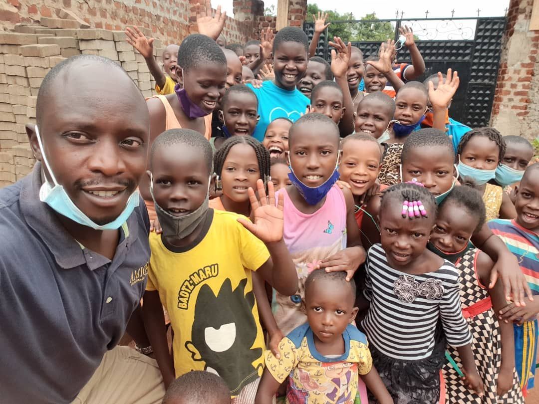 Man with mask smiles, surrounded by many waving children outdoors near a building.