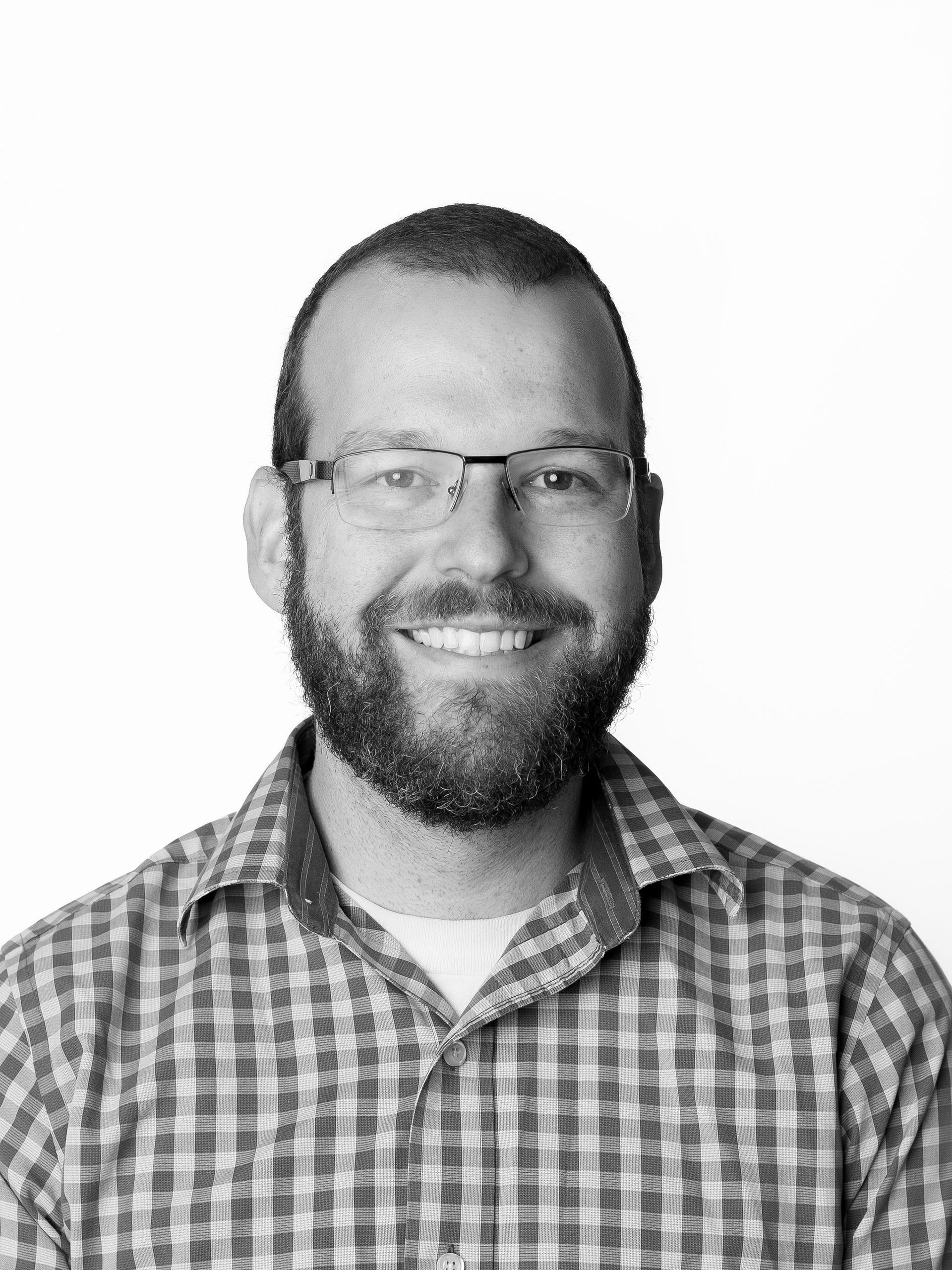A man with a beard and glasses is wearing a plaid shirt and smiling.