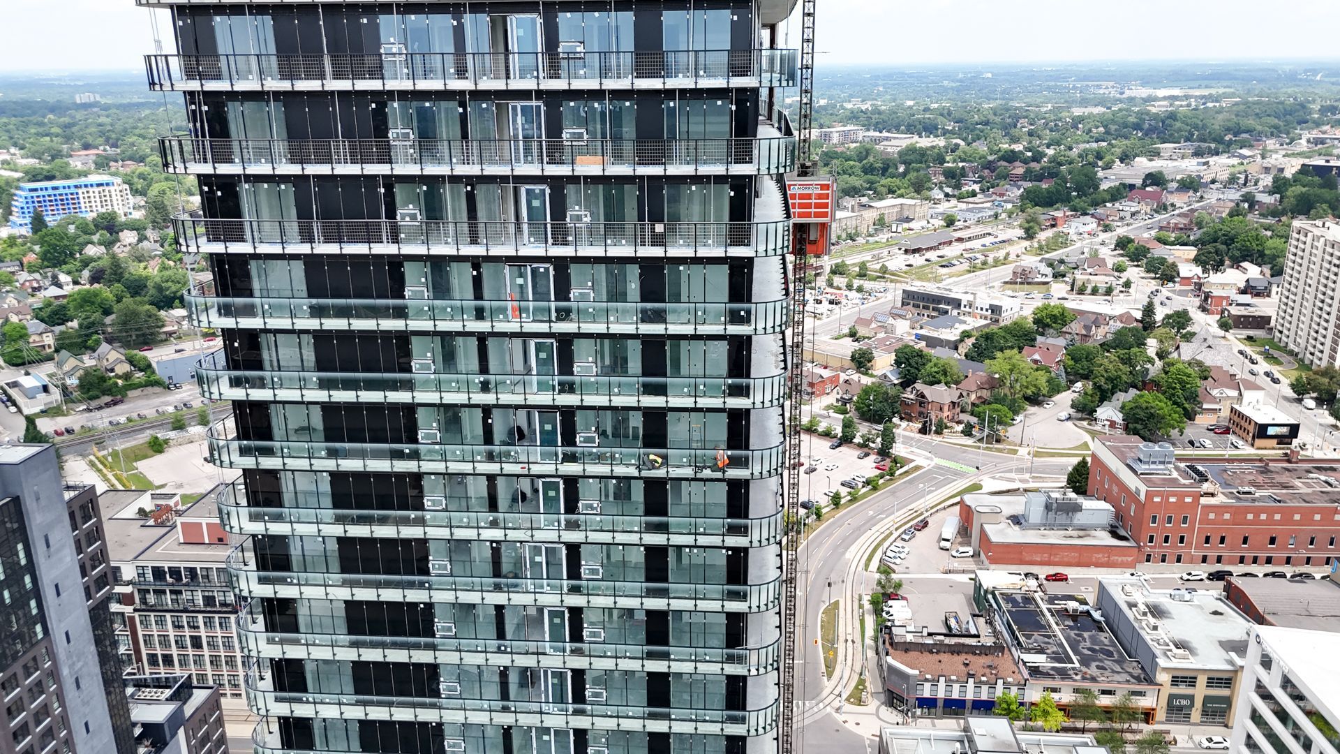 High-rise glass building with balconies, overlooking a city with roads, buildings, and green spaces.