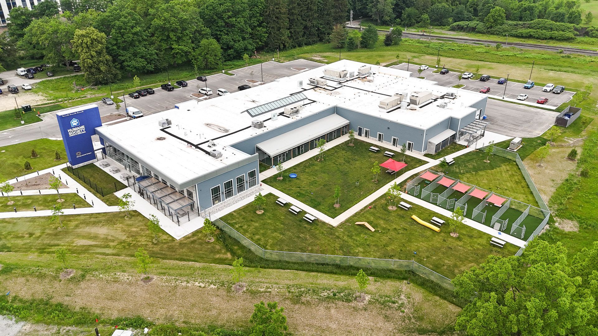 Aerial view of a large gray building with a grassy courtyard, dog runs, and parking lot surrounded by trees.