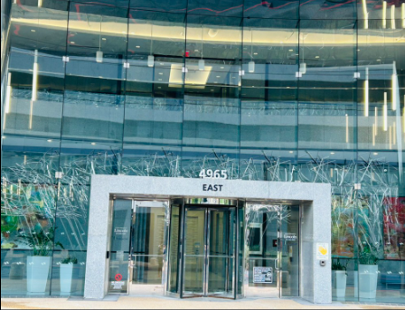 Brown brick office building with large windows, surrounded by trees and a paved walkway.
