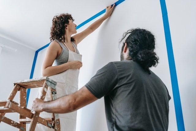 person-standing-on-a-ladder-painting-a-wall-while-another-person-holds-the-ladder