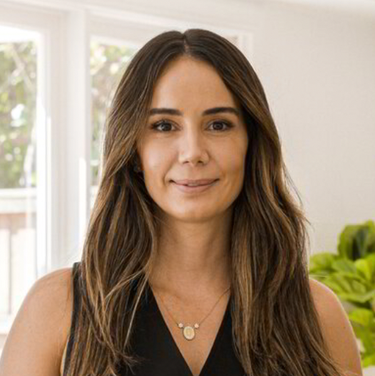 Woman with long brown hair, wearing a black top, smiling in a bright room.