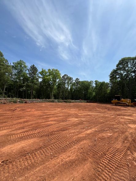 A cleared, reddish-brown construction site under a blue sky, bordered by a treeline. A bulldozer sits in the distance.