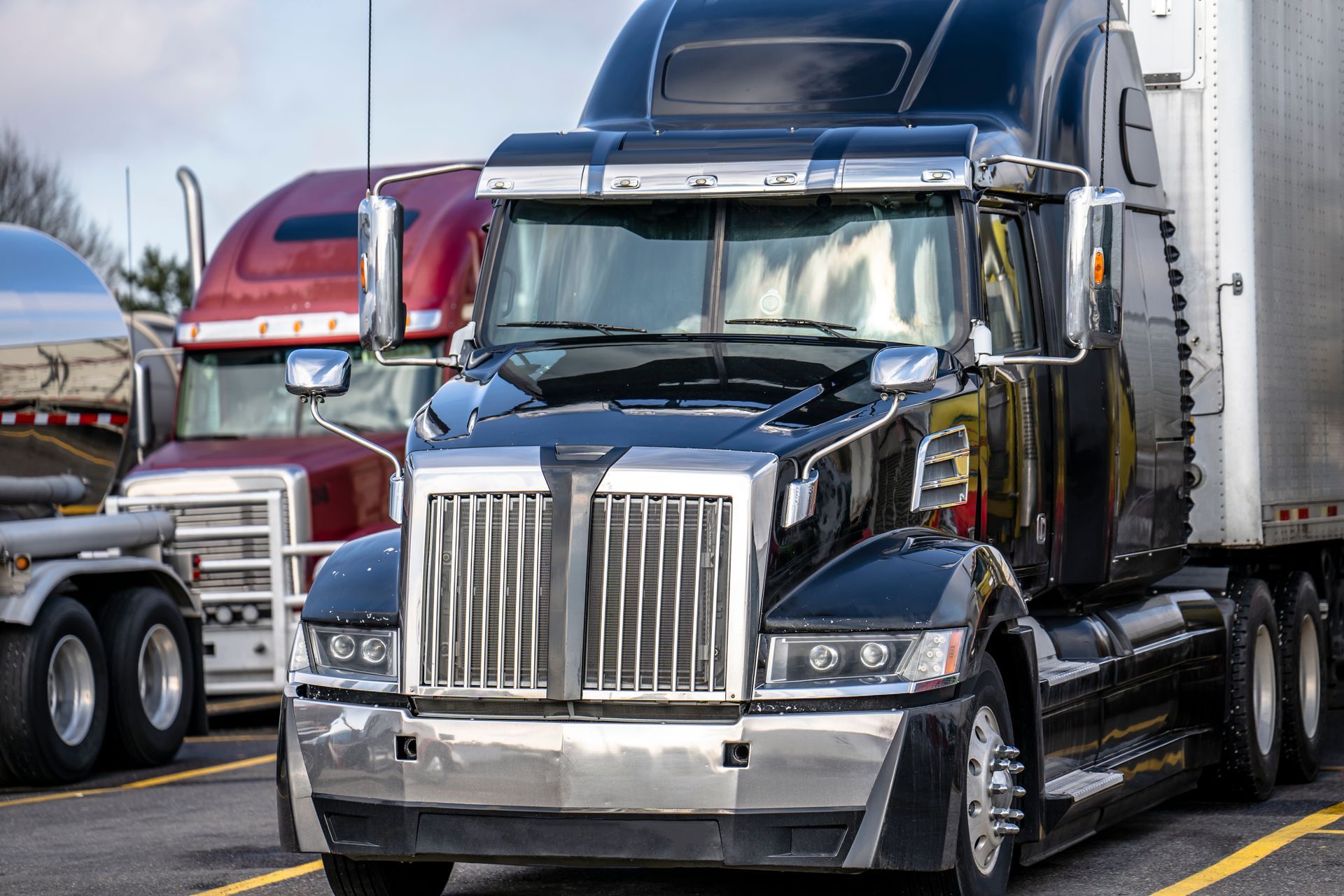 Black semi-truck with chrome grill in a parking lot, with a red truck in the background.
