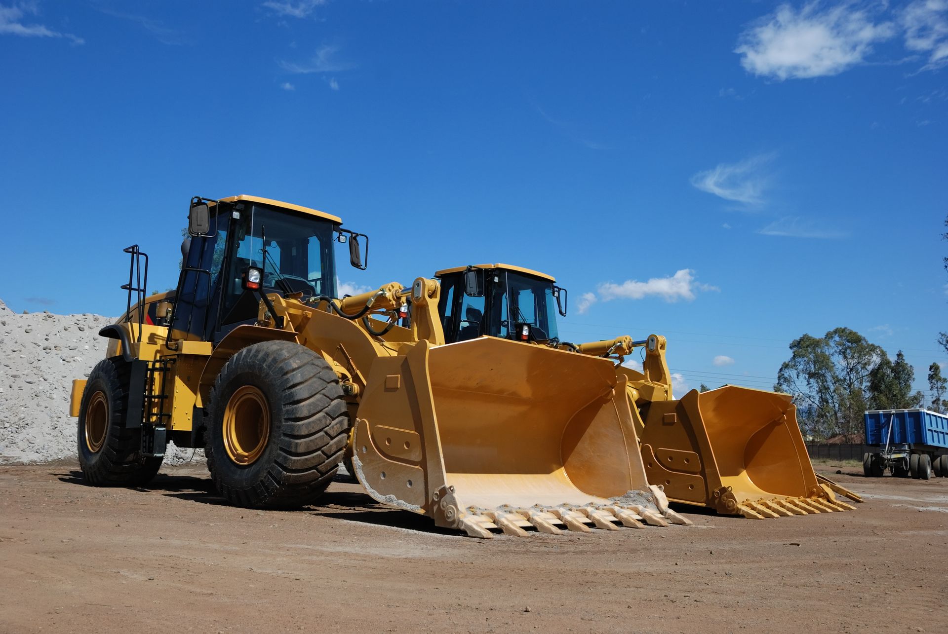 Two yellow front-end loaders parked on dirt under a bright blue sky.