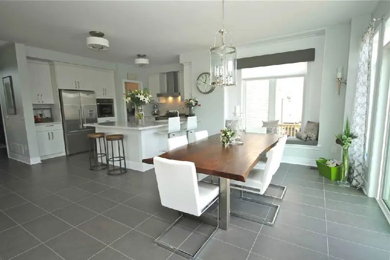 A bright, modern kitchen and dining area featuring gray tile floors, a wooden dining table, and white island stools.