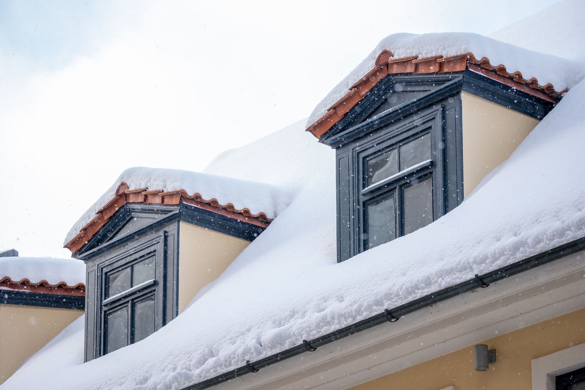 Snow-covered rooftop with dormer windows; light beige walls, dark blue trim, and terracotta roof tiles.