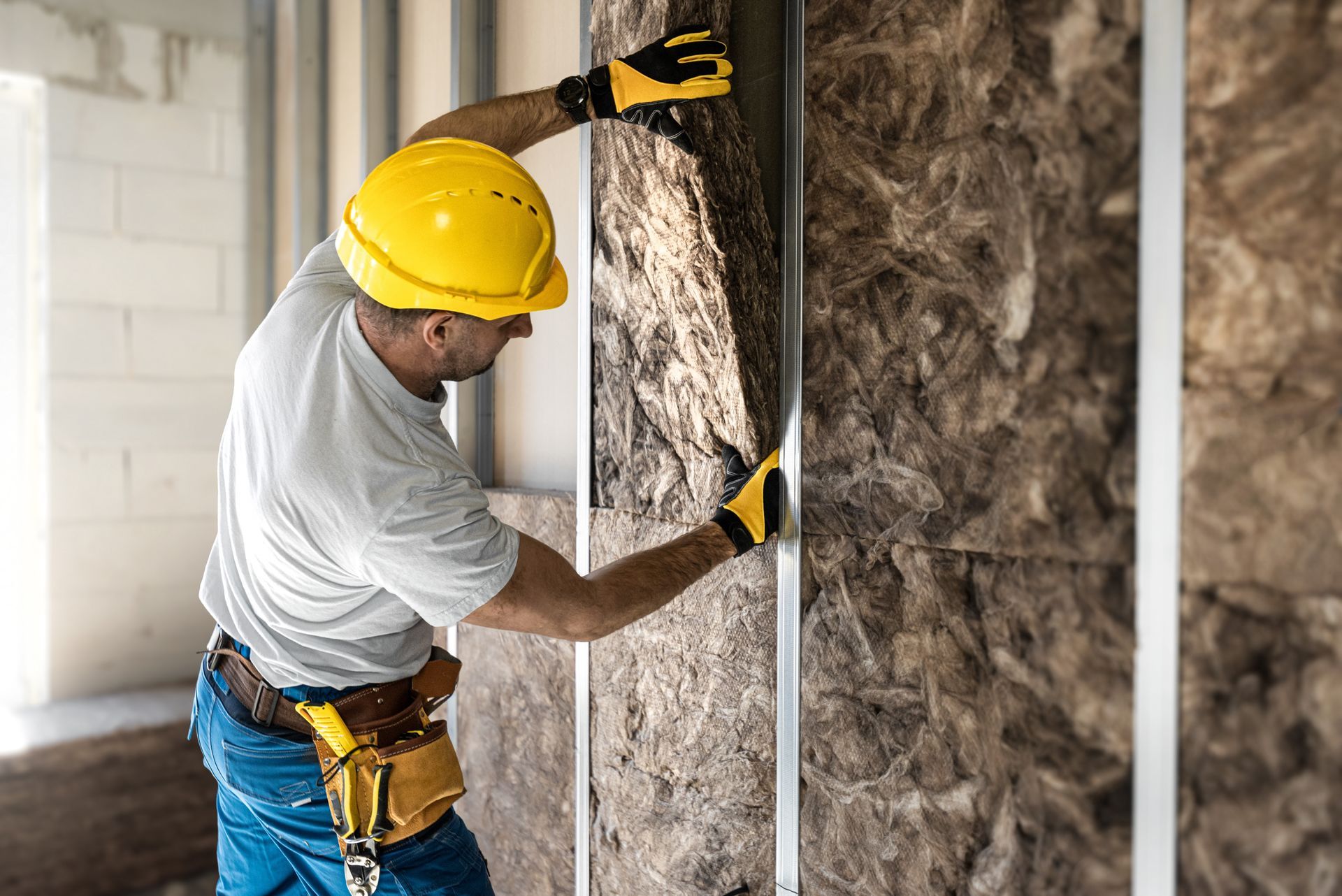 Construction worker in a yellow hard hat installs insulation on a wall, wearing gloves and a tool belt.