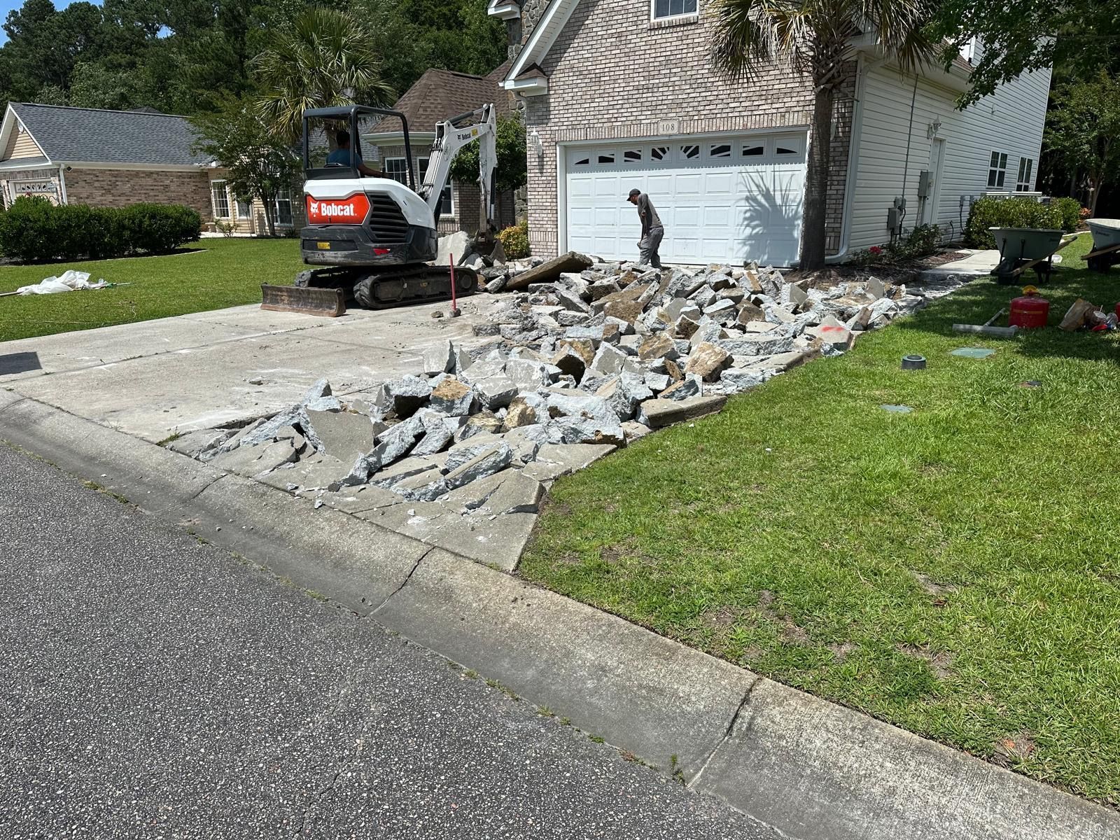 A small excavator is demolishing a concrete driveway in front of a house.