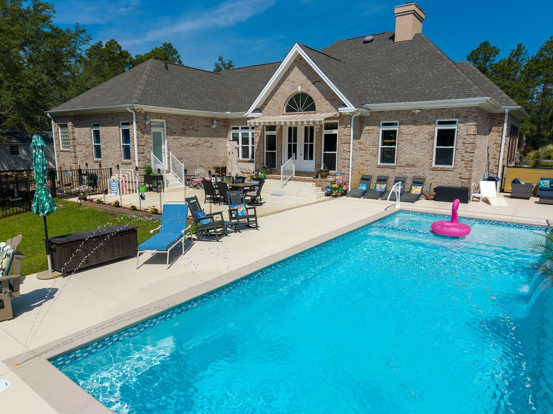 Backyard patio with a large blue pool beside a stone house under a clear sky