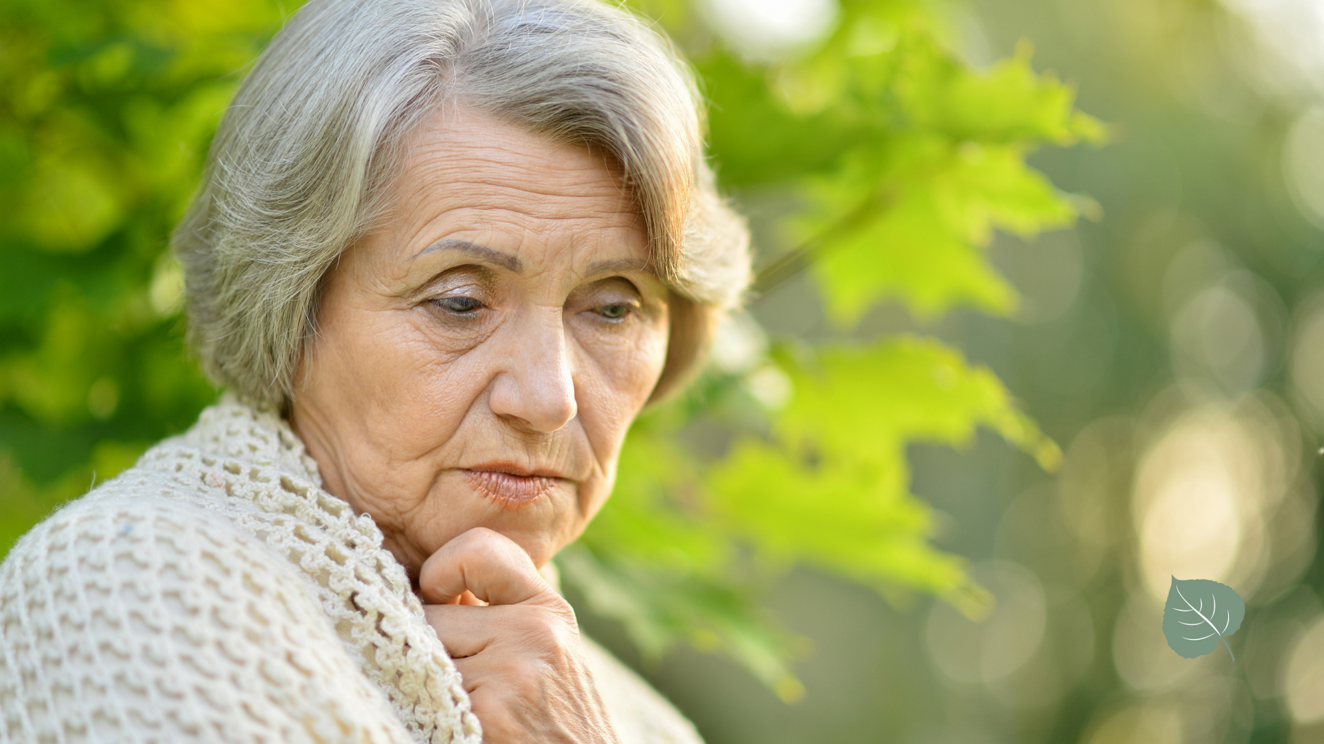 An elderly woman is standing in front of a tree with her hand on her chin.