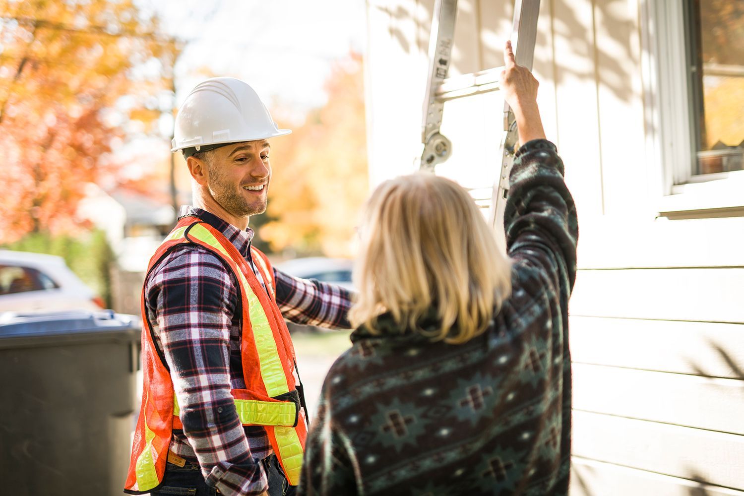 A contractor with a hard hat and safety vest is talking with a customer as she points up at her roof.