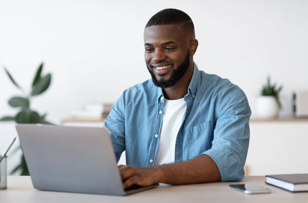 Man smiling while working on laptop at a desk. Indoors, natural light.