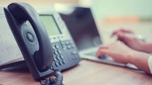 A person is typing on a laptop next to a telephone on a desk.