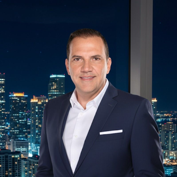 Man in a blue suit smiling, standing by a window at night overlooking a city skyline.