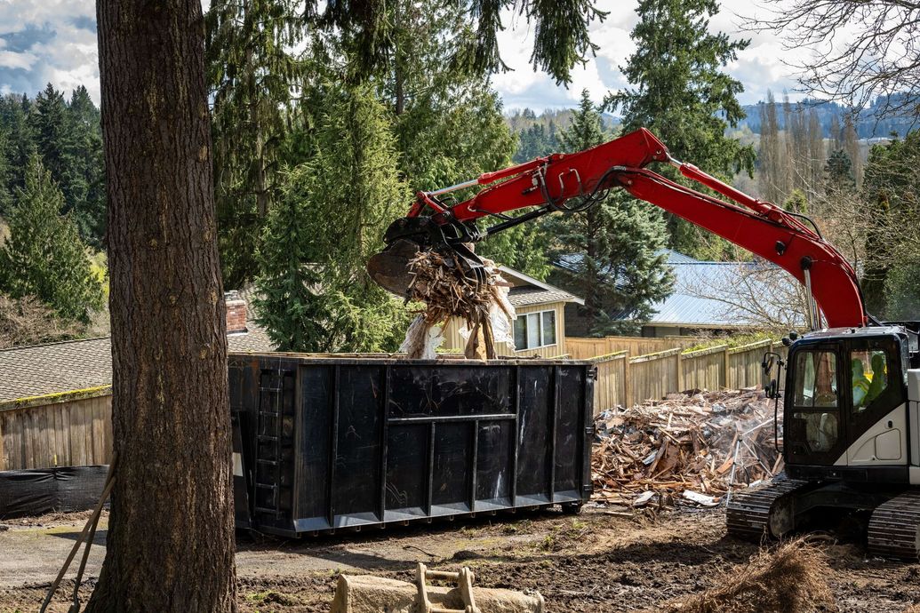 Red excavator dumping debris into a black dumpster in a wooded yard beside a lake