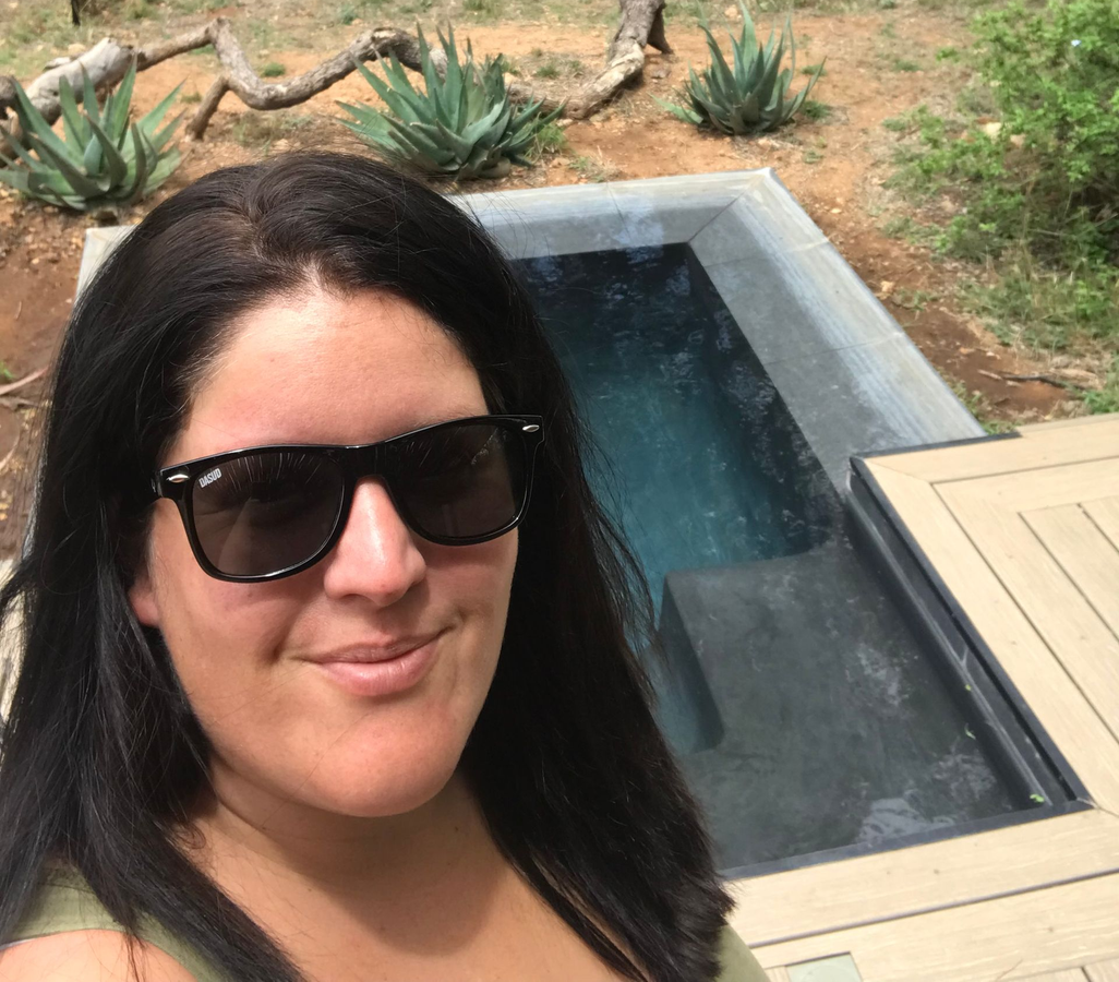 Woman in sunglasses smiles near a small, rectangular pool. Agave plants and a wooden deck are in the background.