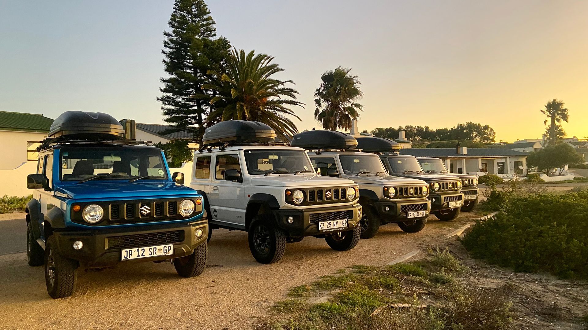 Lineup of five SUVs with roof cargo carriers on a dirt road, at sunset.