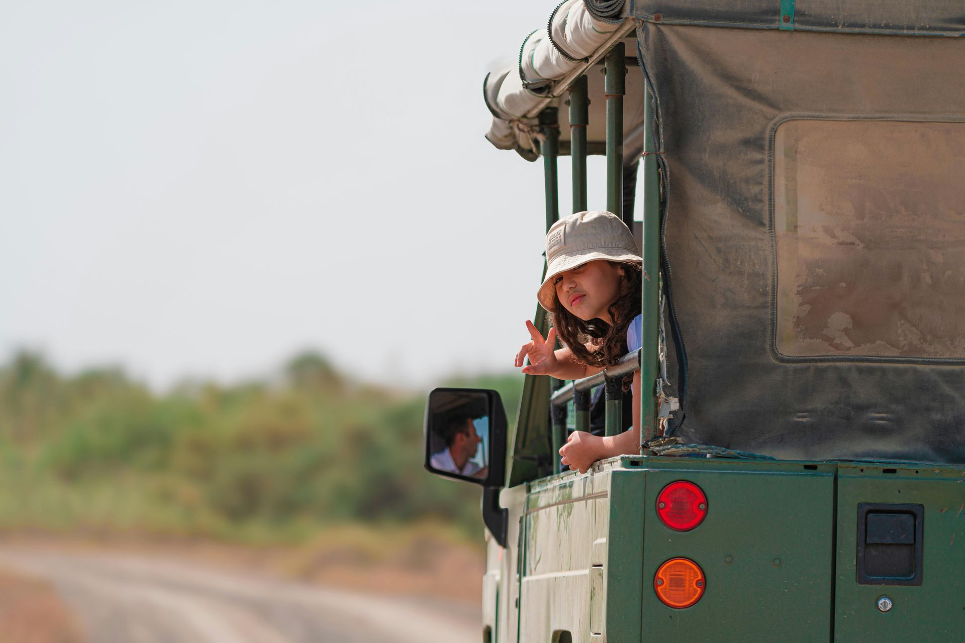 Girl in a safari vehicle gives the peace sign, looking out at a dirt road.