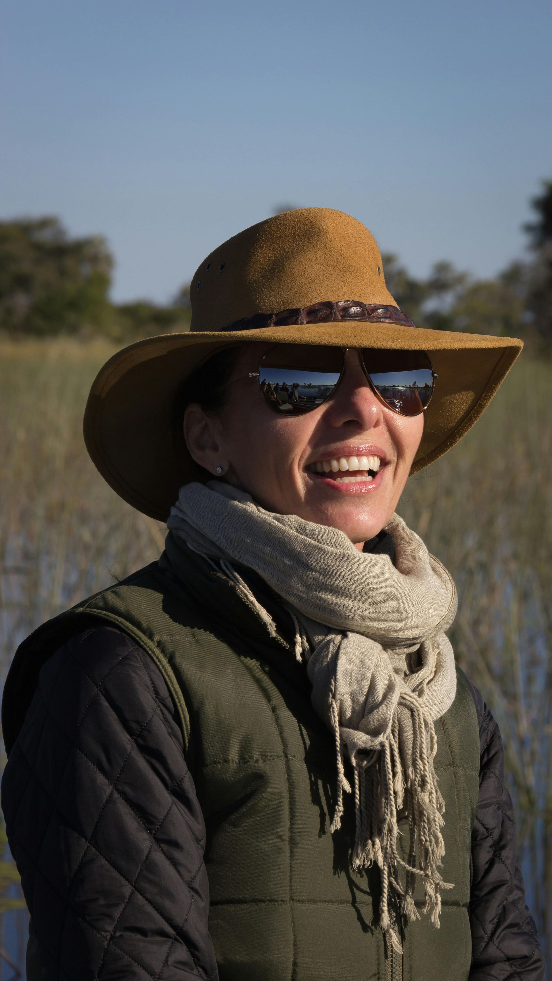 Woman wearing a hat and sunglasses, smiling. She has a scarf and a green vest outdoors.