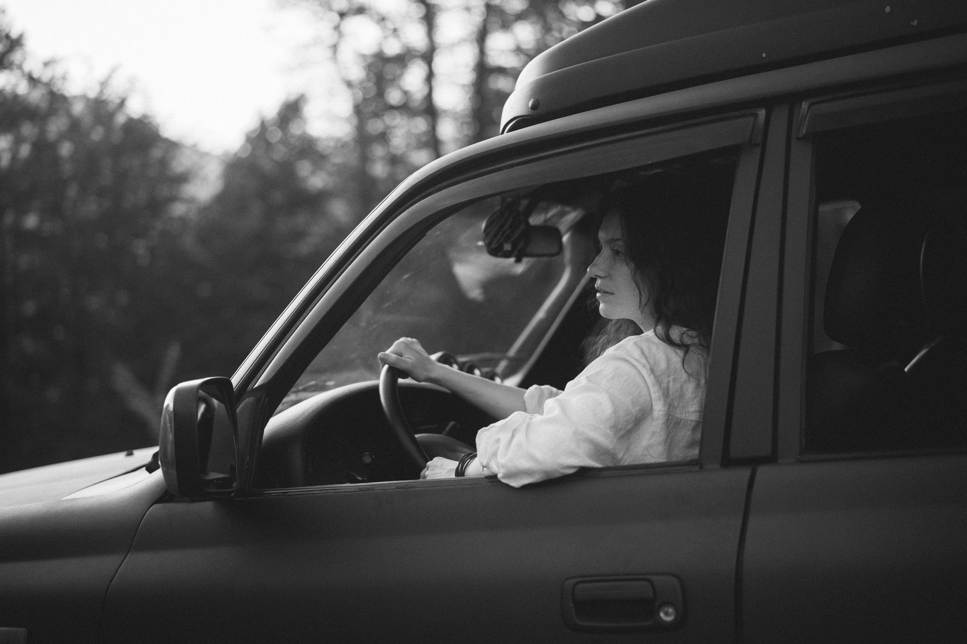 Woman driving a dark-colored SUV, looking ahead, with a forest backdrop, in black and white.