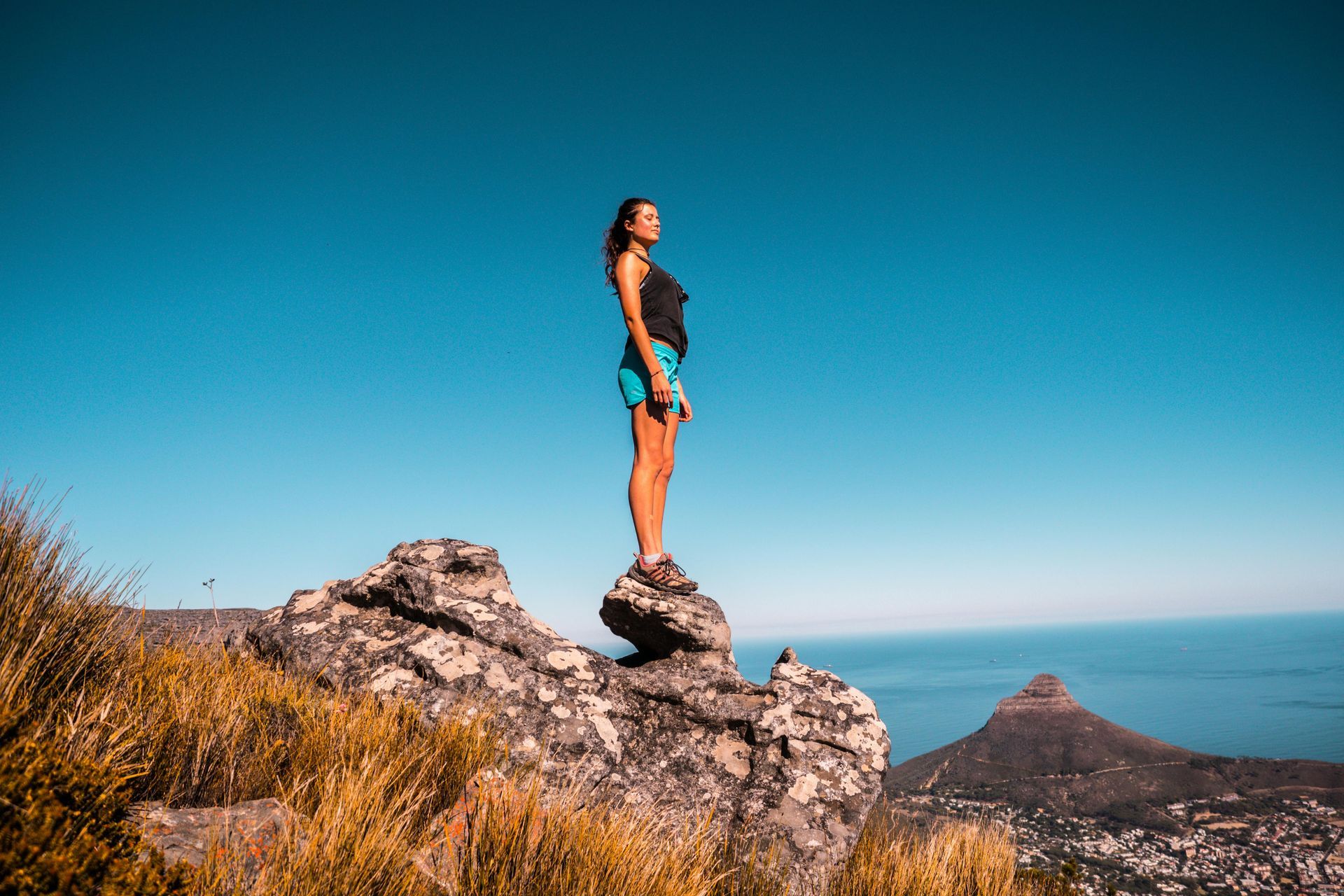 Woman standing on a rock, looking up with arms at side. Blue sky and ocean in background.