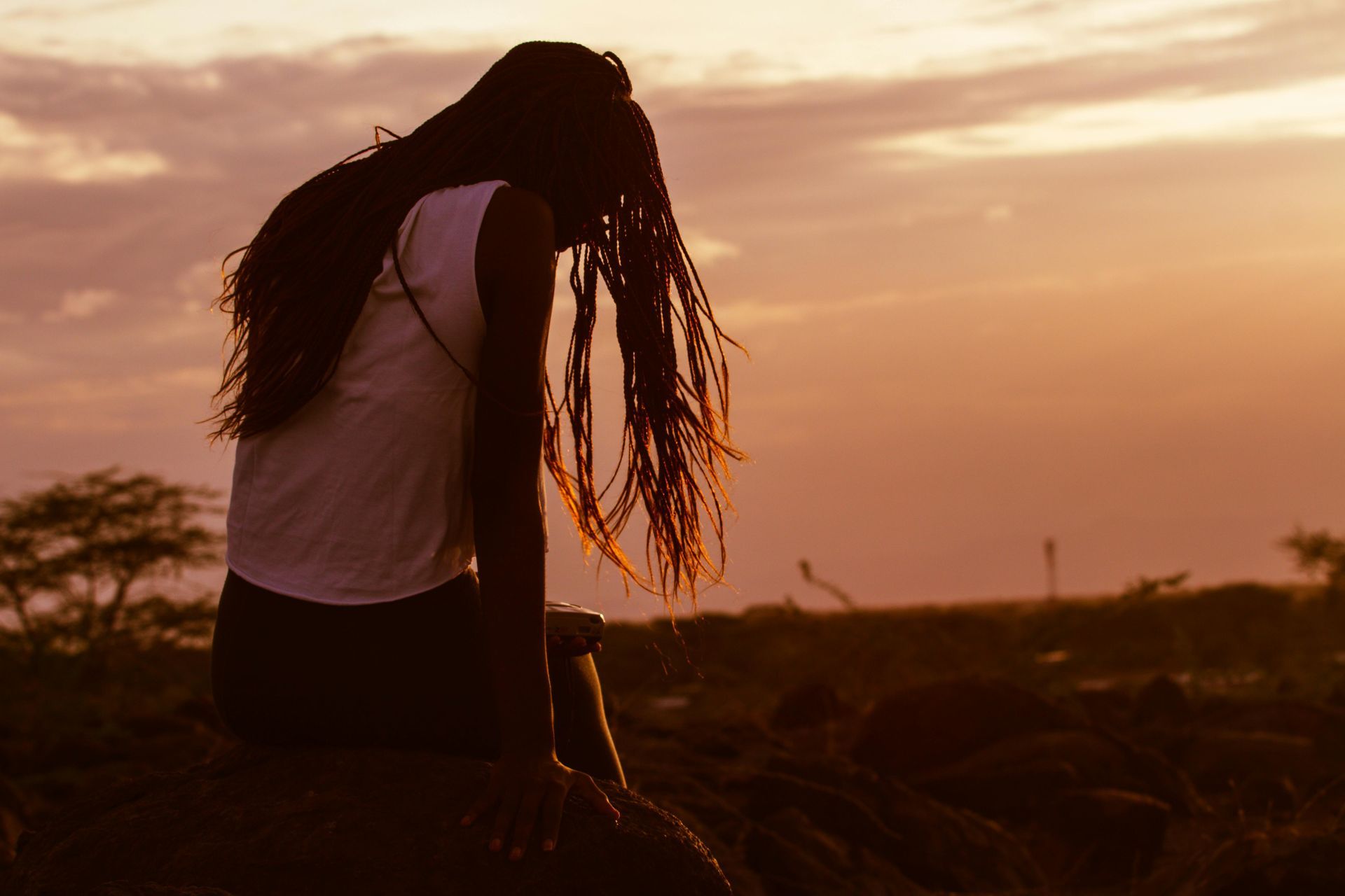 Person with long hair sits on a rock, silhouetted against a golden sunset.