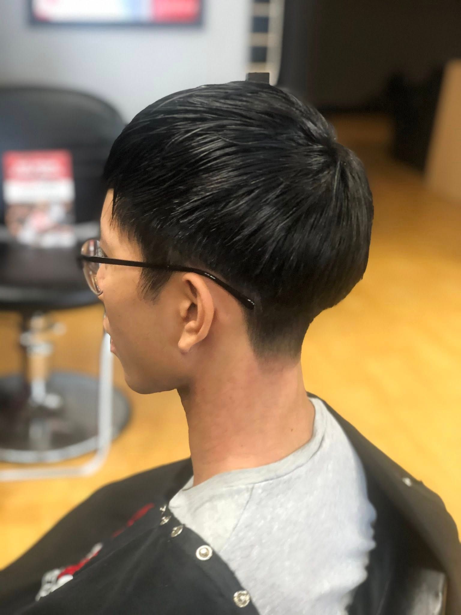 Side profile of a person with glasses and a short dark haircut in a salon chair, wearing a gray T-shirt and cape