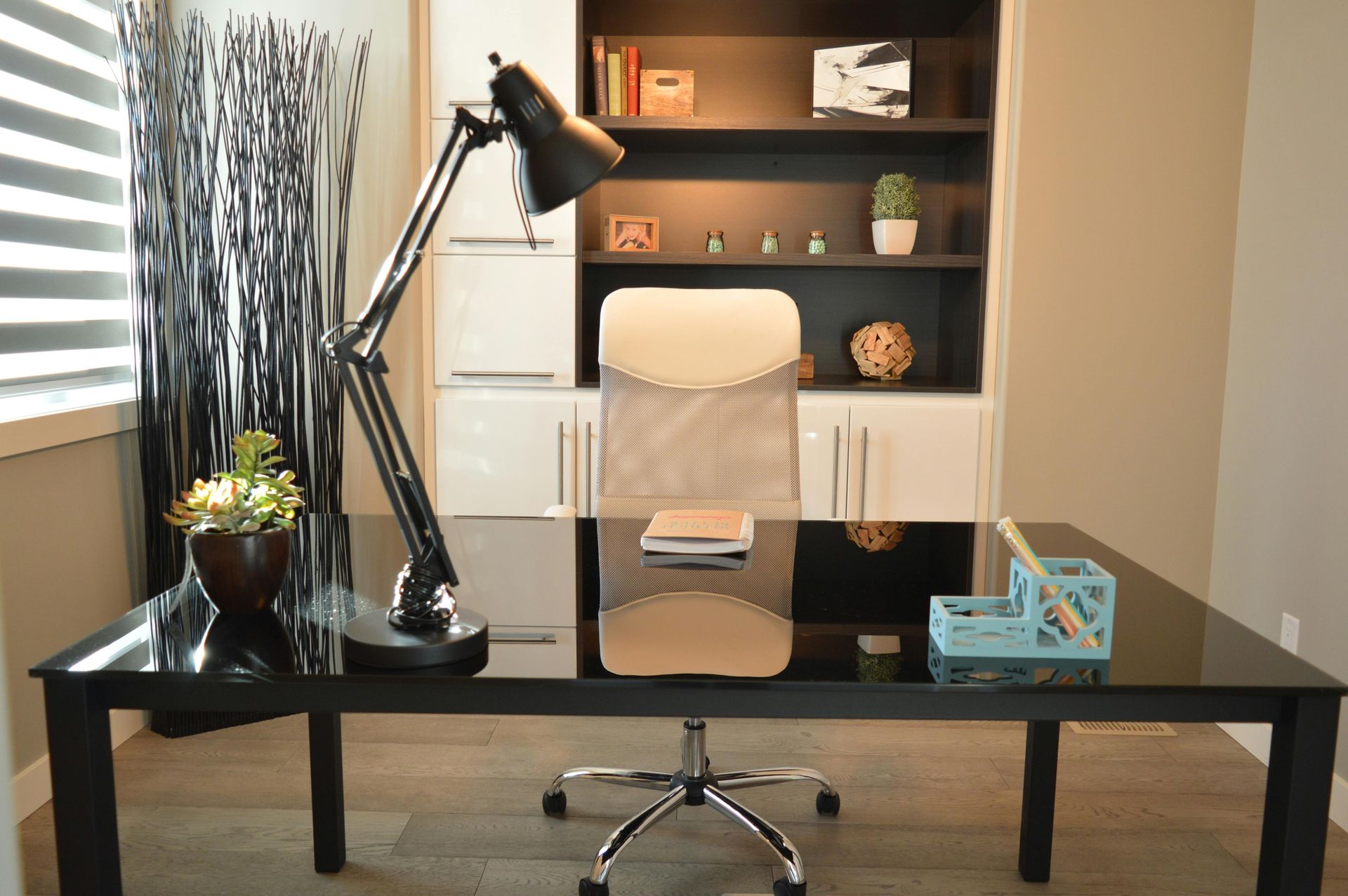 A modern home office with a black glass desk, a desk lamp, an office chair, and shelves decorated with small items.