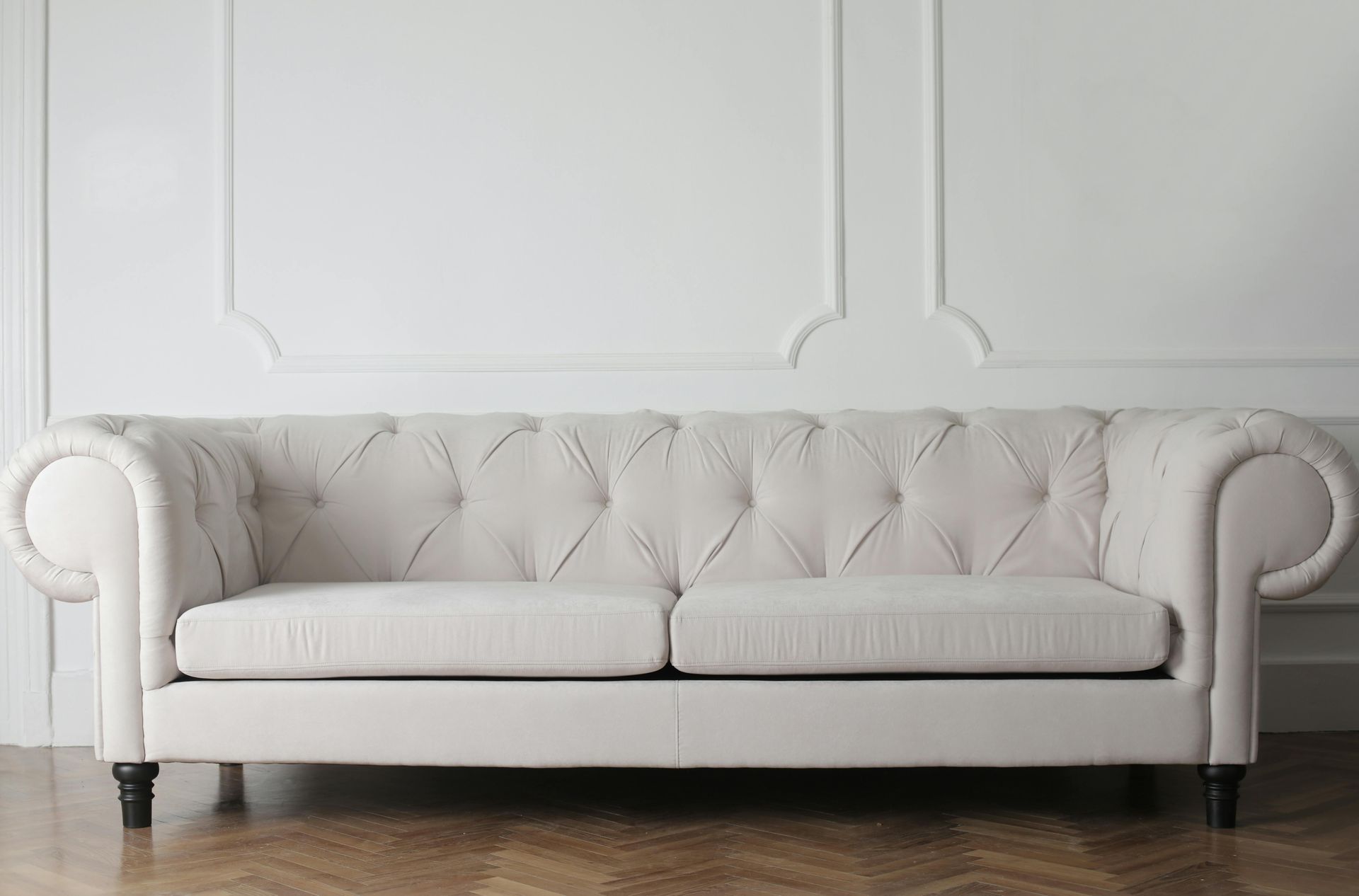 Off-white, button-tufted Chesterfield sofa against a white paneled wall with a dark wood herringbone floor.
