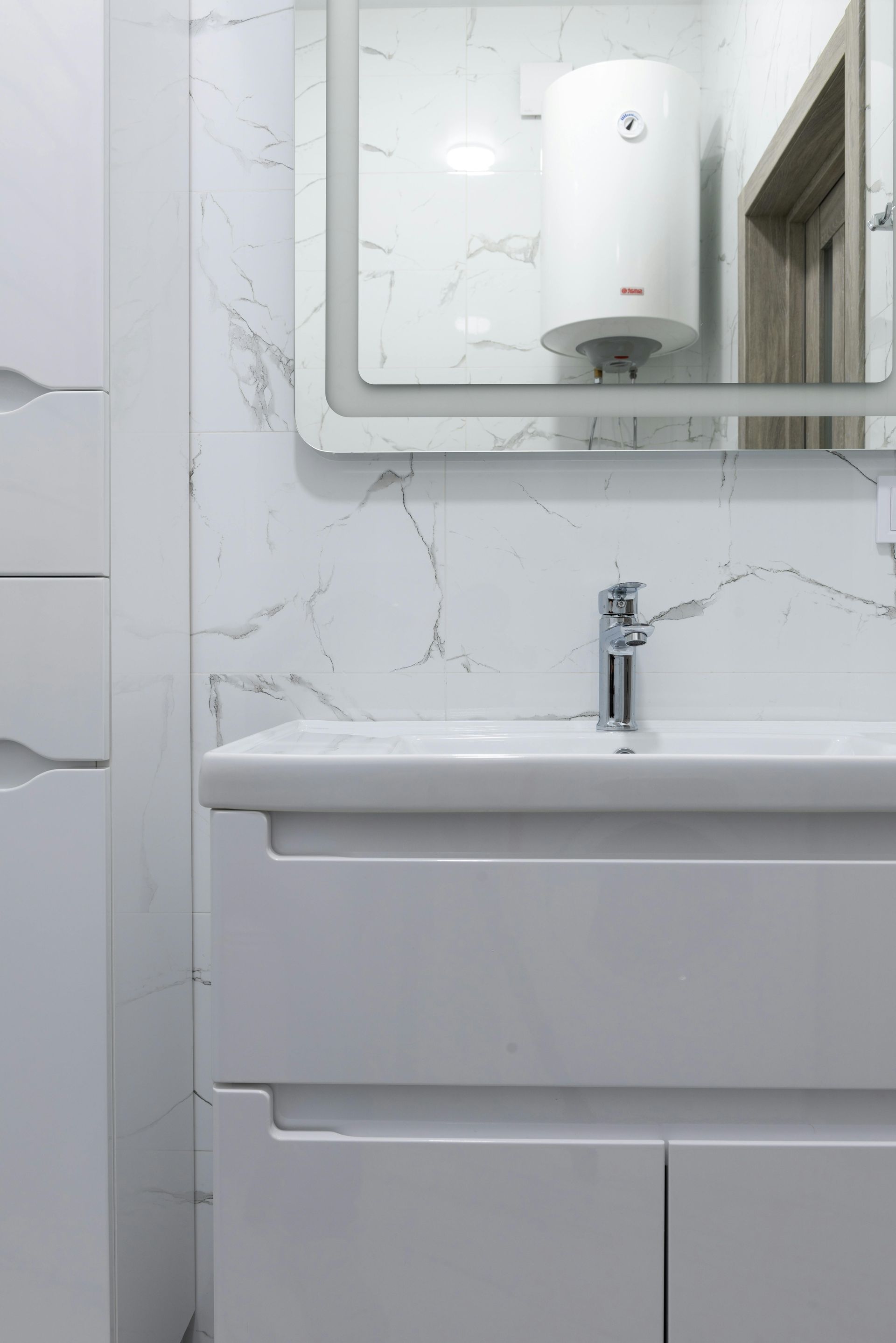 White bathroom with a sink, mirror, and water heater; marble-patterned wall.