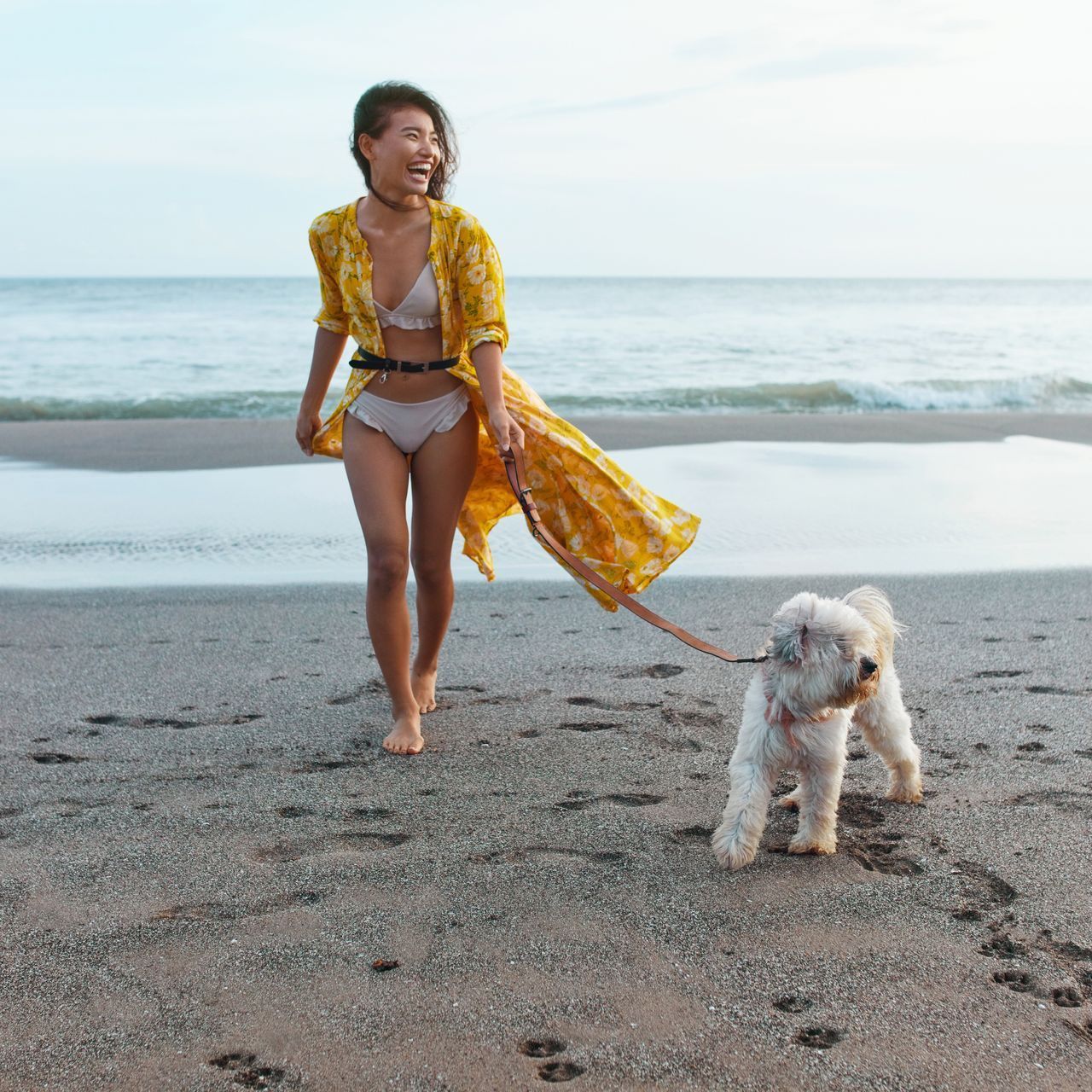 A woman in a bikini is walking a small dog on the beach