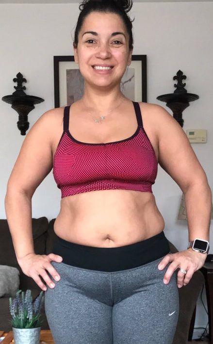 A woman in a red sports bra and grey pants is standing in a living room with her hands on her hips.