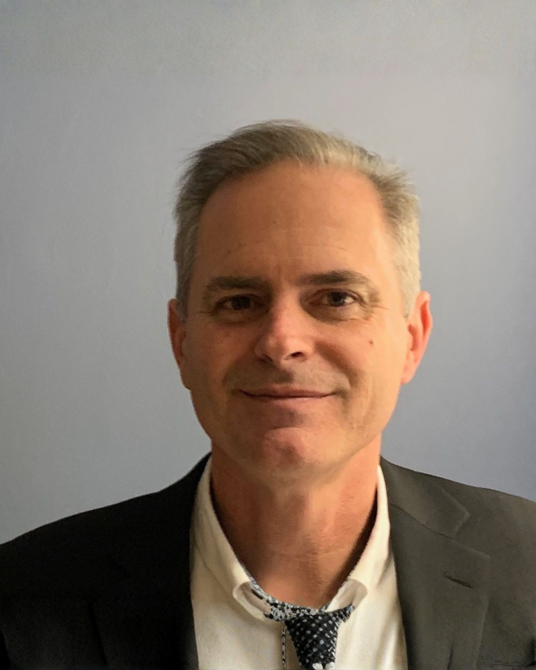 Smiling man in a dark blazer and loosened tie against a plain gray background