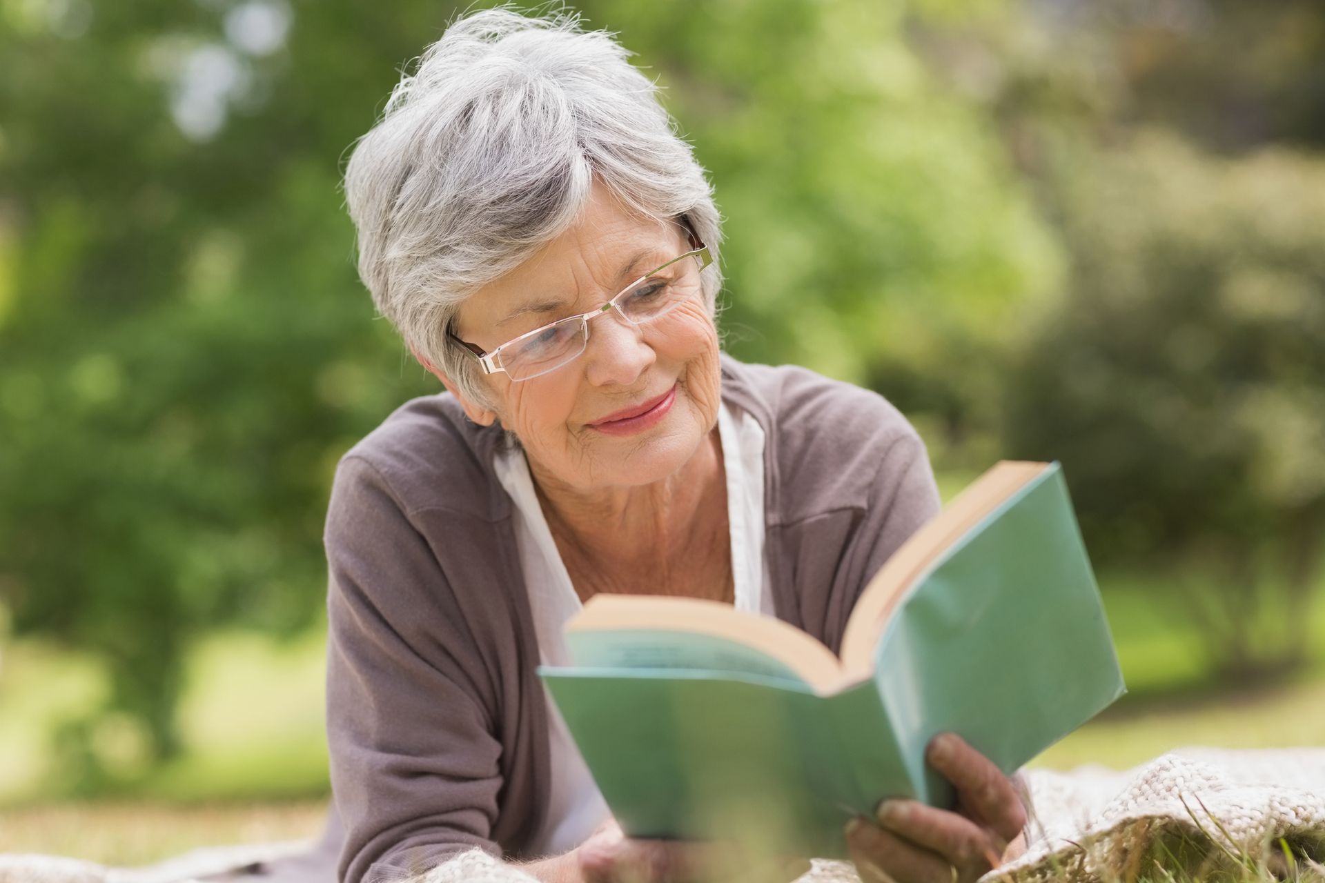 an elderly woman is laying on a blanket reading a book .