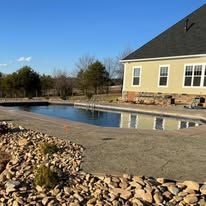 Exterior view of a house with a pool. The water reflects the building. A rock border surrounds the pool deck.