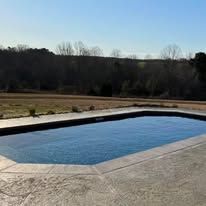 Rectangular outdoor swimming pool with concrete surround, in a grassy field on a sunny day.