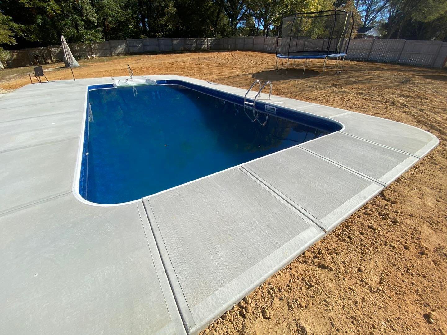 Concrete-surrounded pool filled with blue water. Newly built in a backyard with dirt and a trampoline.