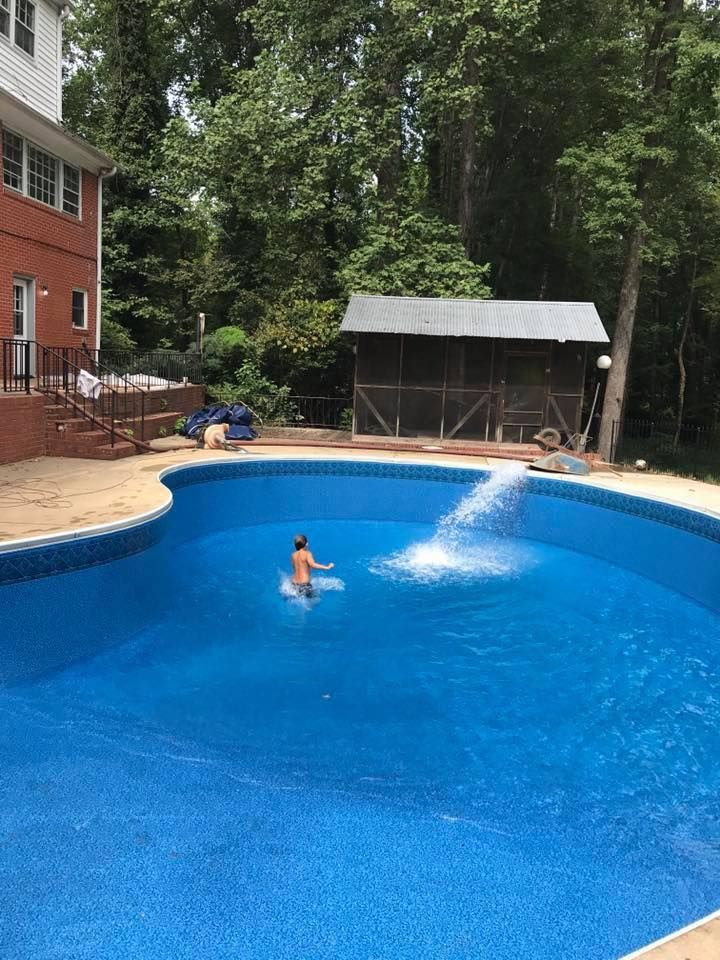 Child playing in a blue swimming pool with water fountain, house and trees in the background.