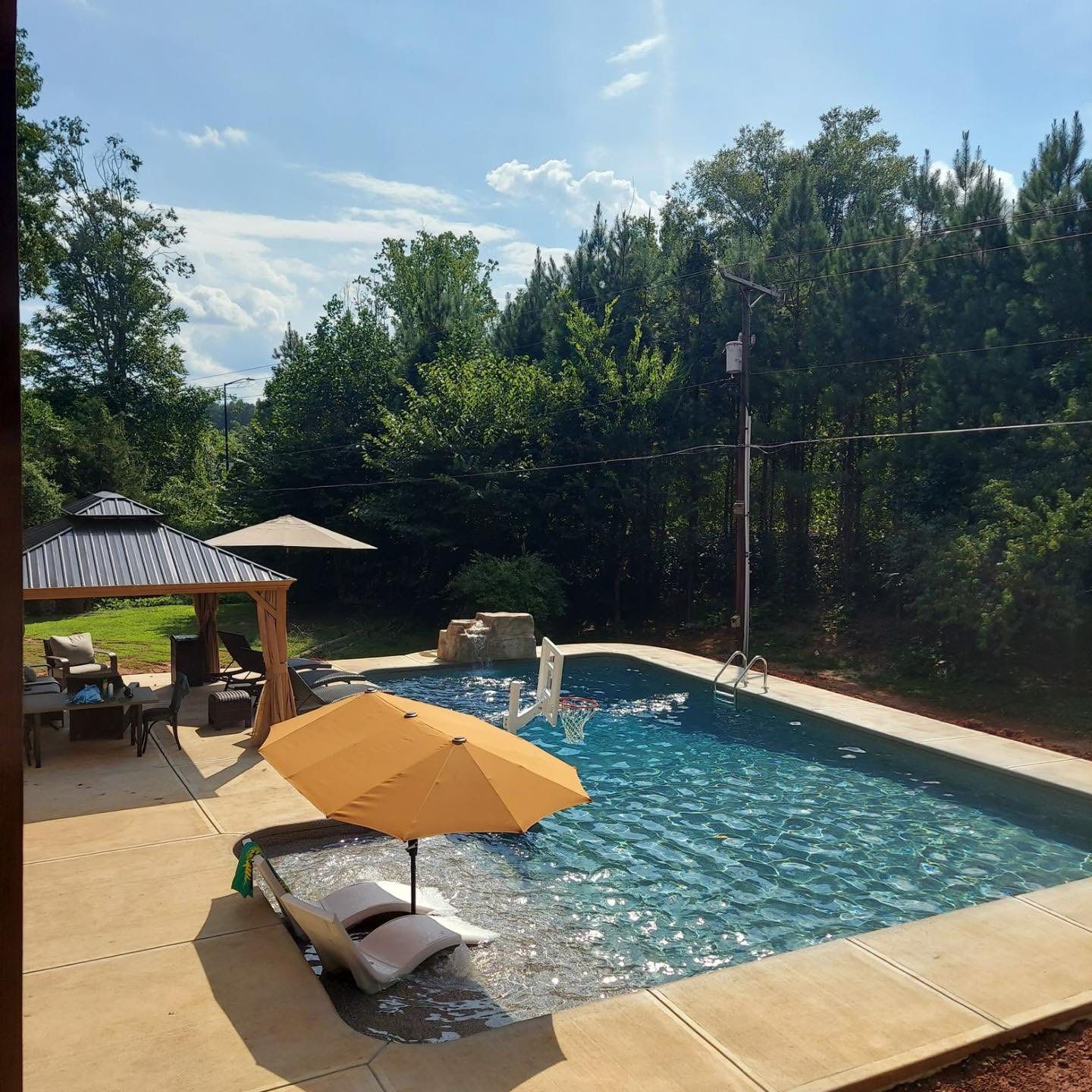 Poolside scene with a pool, gazebo, and trees under a sunny sky.