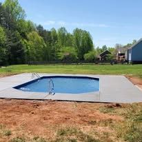 A rectangular swimming pool surrounded by concrete, in a grassy yard, with trees in the background under a blue sky.