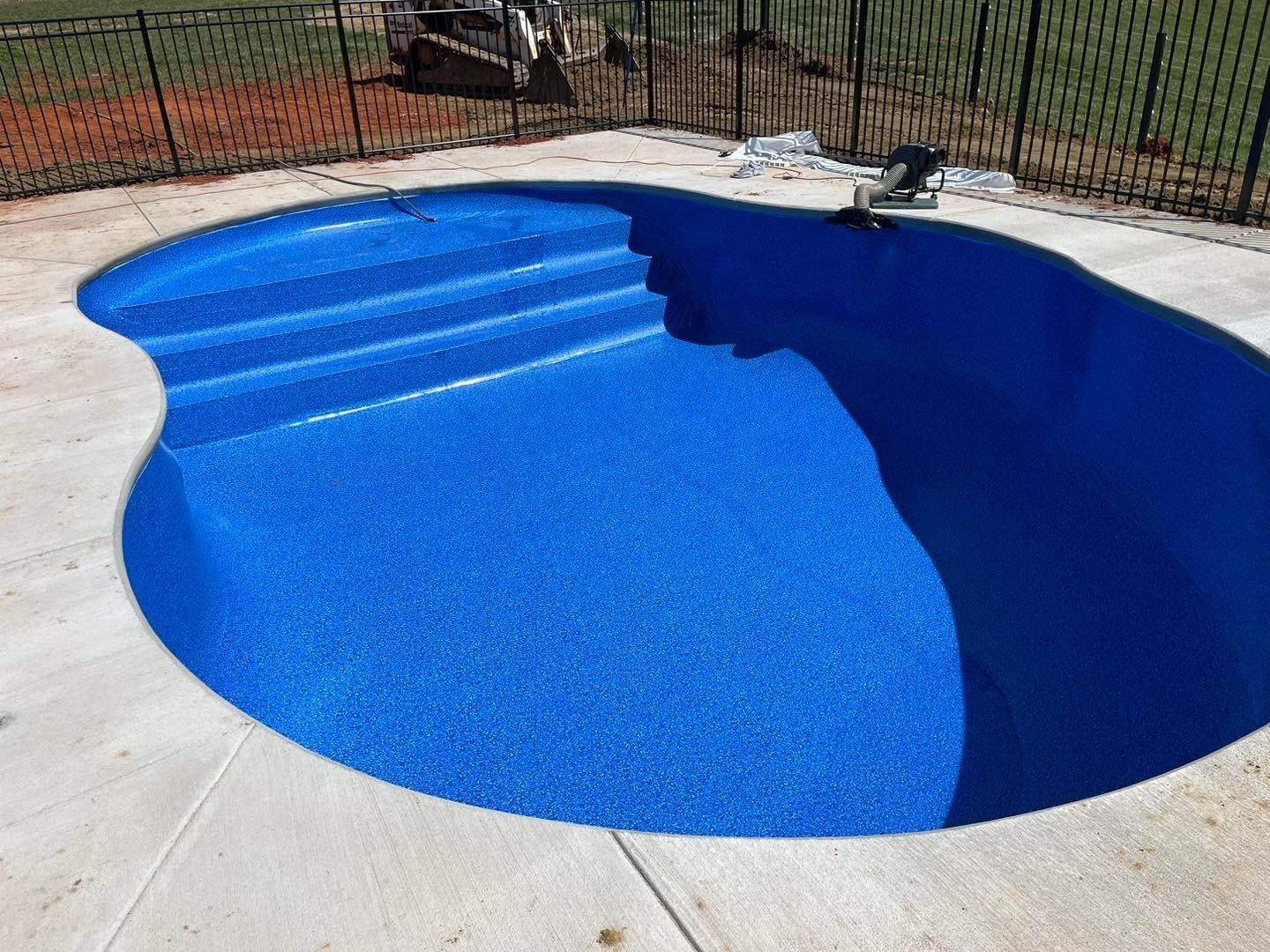Blue, finished swimming pool with built-in steps, surrounded by concrete and a black fence.