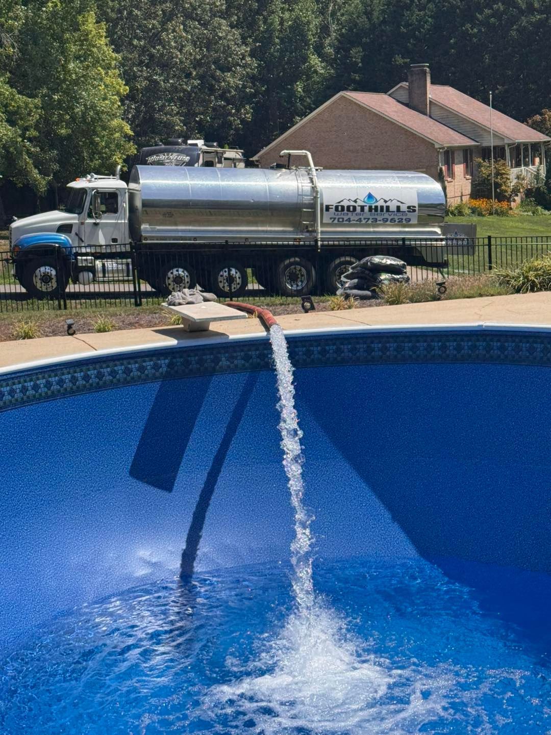 A silver tanker truck filling a blue swimming pool with water.