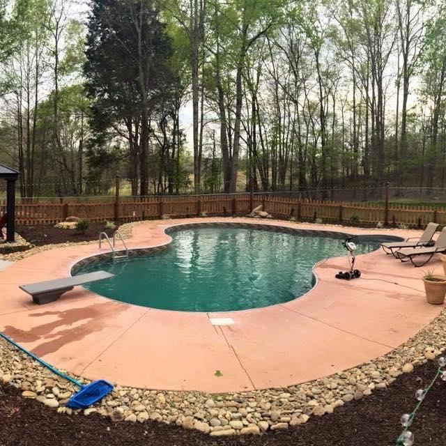 Swimming pool with concrete deck, diving board, and lounge chairs surrounded by a wooden fence and trees.