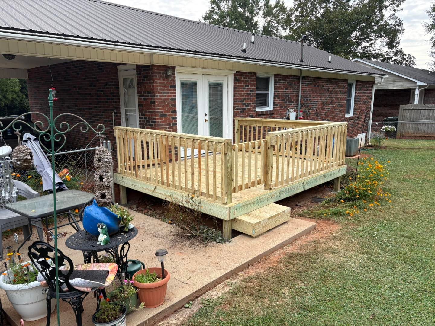 Wooden deck attached to a brick house with steps, surrounded by a yard with plants and patio furniture.
