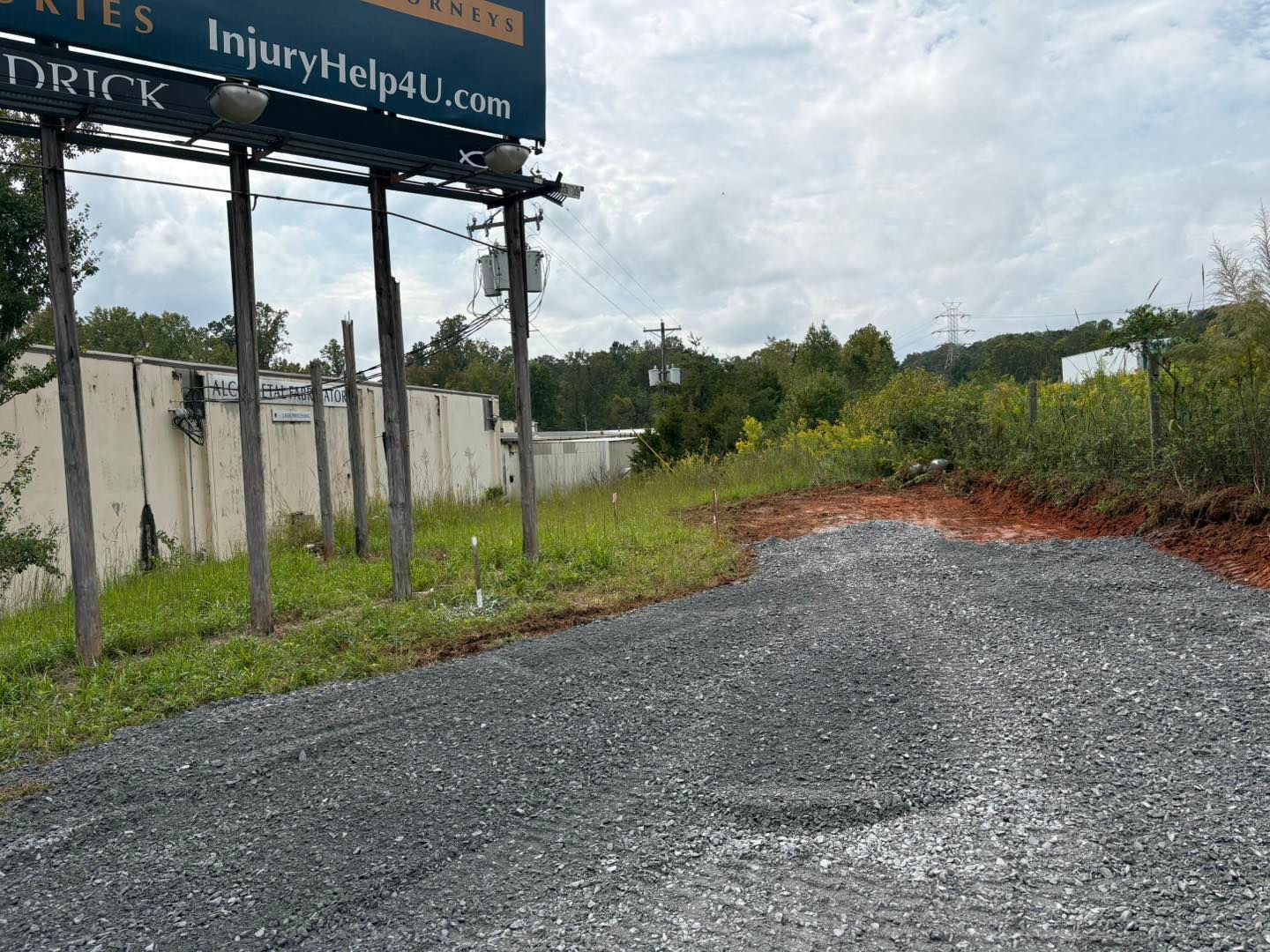 Gravel driveway leads toward grassy area with a billboard and utility poles on a cloudy day.